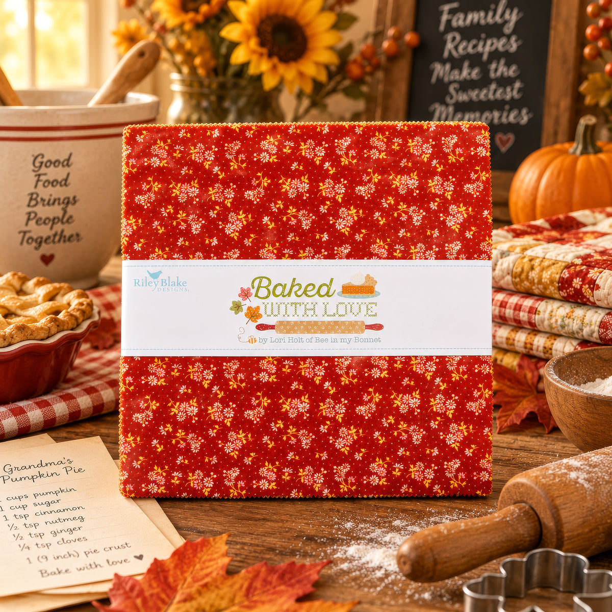 A folded fabric bundle with a red floral print and a white paper band reading 'Baked with Love' is displayed on a rustic wooden table surrounded by autumn-themed quilting props, including a pie, sunflowers, a rolling pin, and cookie cutters.
