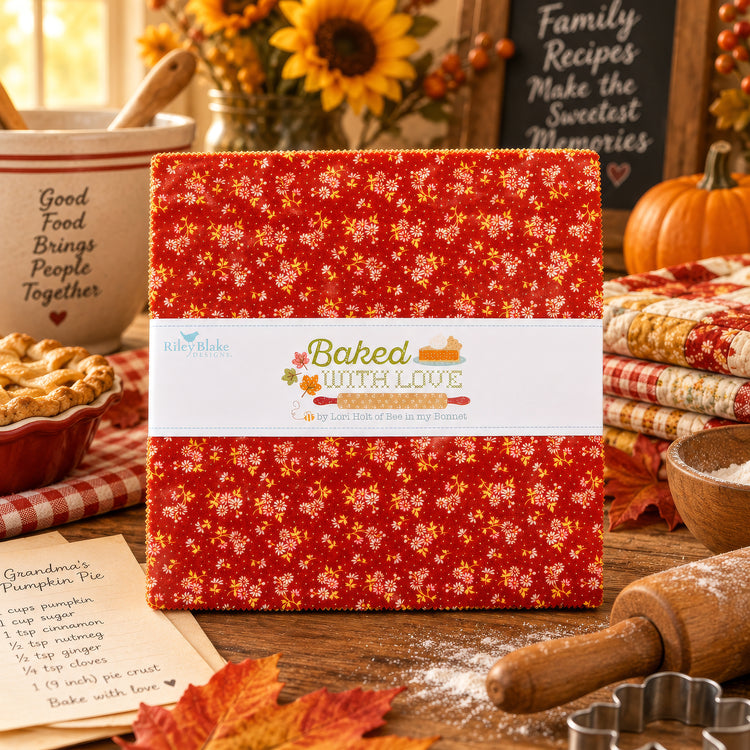 A folded fabric bundle with a red floral print and a white paper band reading 'Baked with Love' is displayed on a rustic wooden table surrounded by autumn-themed quilting props, including a pie, sunflowers, a rolling pin, and cookie cutters.