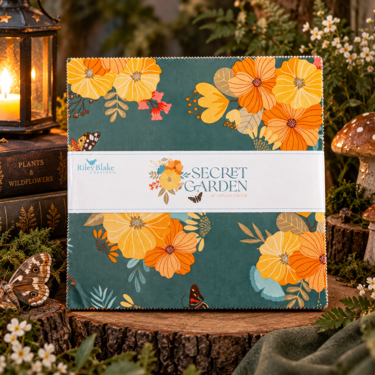 A square fabric sample or quilt block wrapped with a white paper band reading 'Riley Blake Fabrics' and 'Secret Garden', displayed on a rustic wood stump with flowers, candles, and mushrooms in the background.