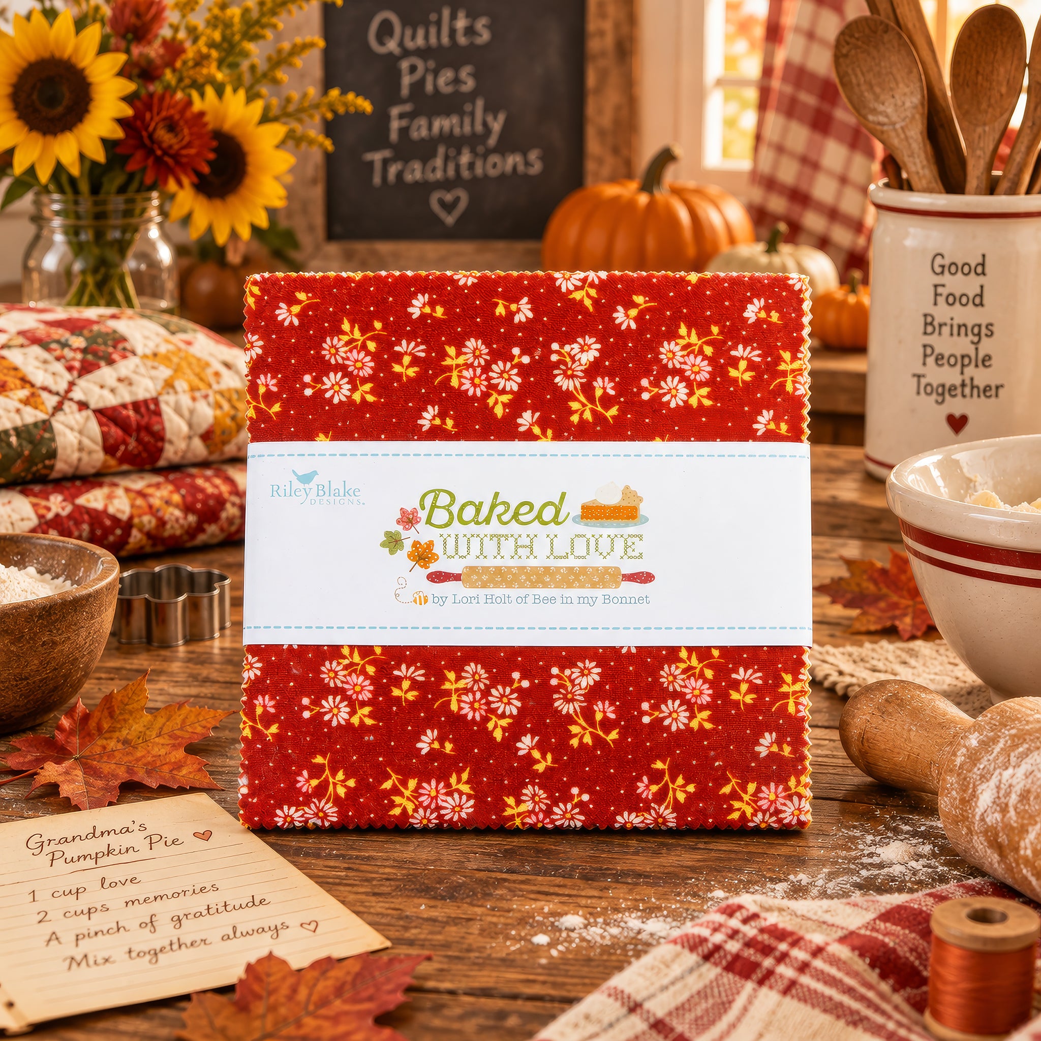 A quilt fabric bundle titled 'Baked with Love' featuring a red and orange floral pattern, displayed on a rustic wooden table surrounded by autumn quilting props like pumpkins, sunflowers, and baking utensils.