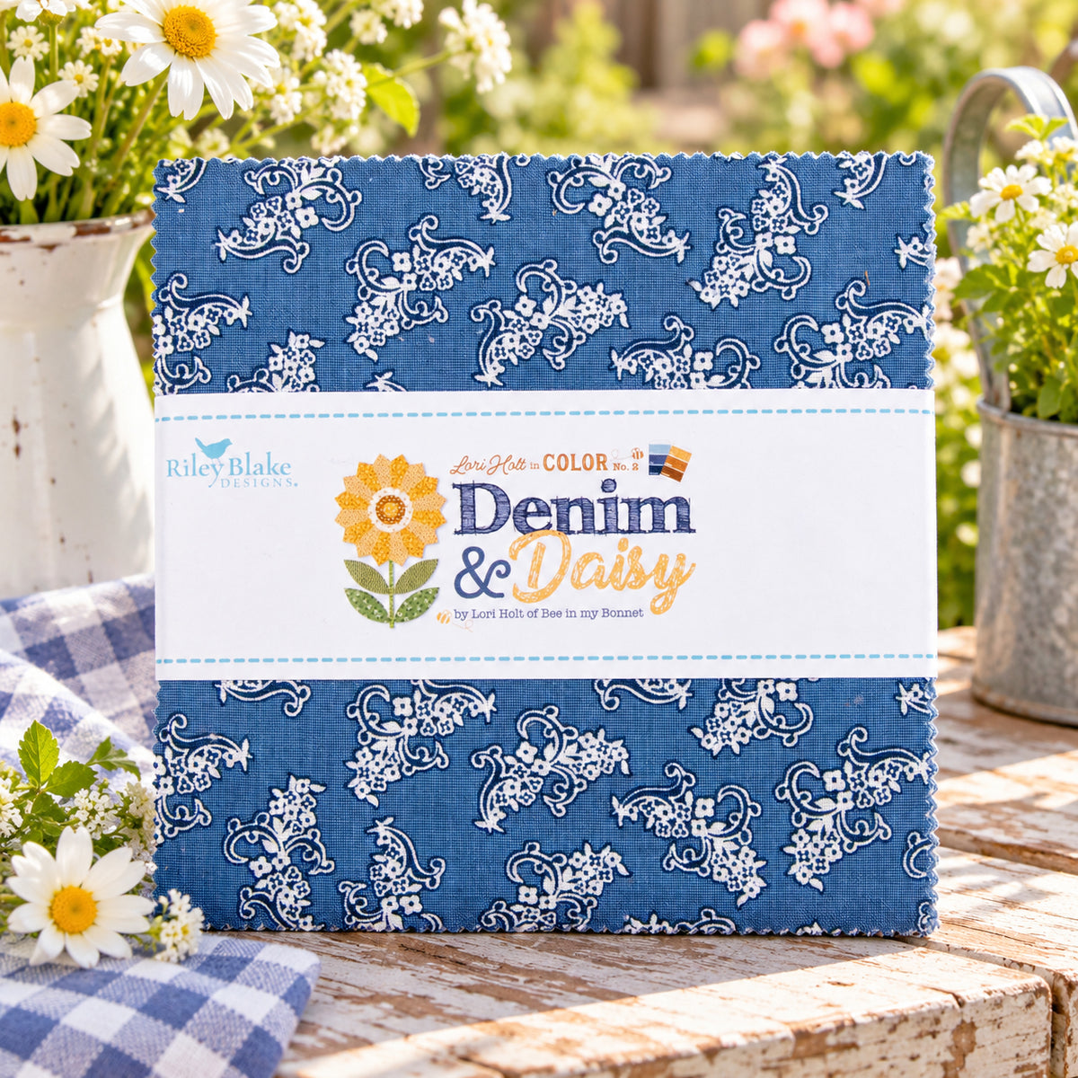 A fabric bundle wrapped with a white paper label placed on a wooden surface with flowers and a gingham cloth in a garden setting.