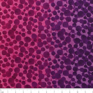 Bumbleberries Pinky Purple fabric with irregular dots in shades of magenta, plum, and violet over a pink-to-purple gradient background with a ruler for scale. 