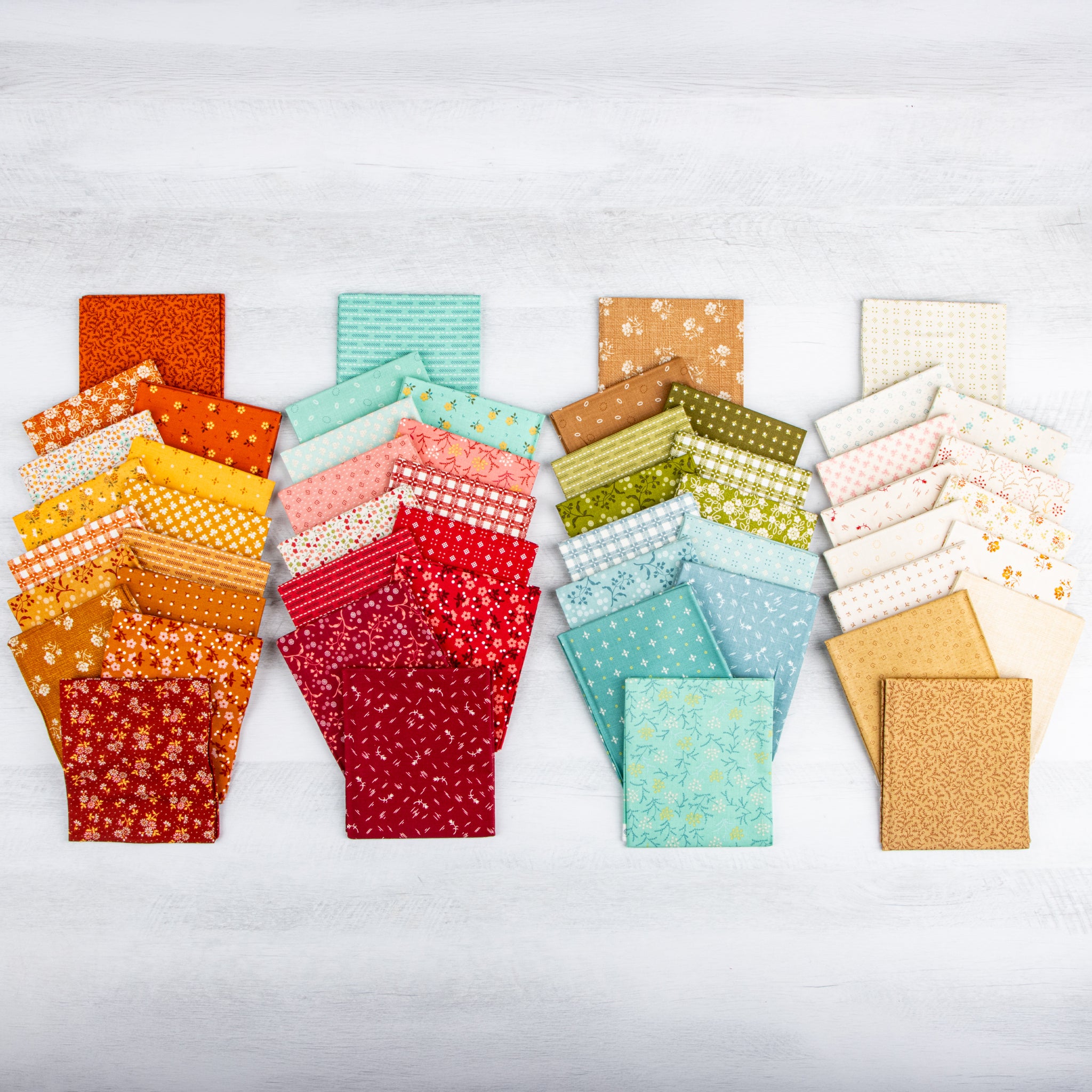 A collection of folded fabric swatches arranged in four vertical groups on a light gray wood background. The fabrics feature various patterns including florals, stripes, polka dots, and plaid in a wide range of colors including orange, yellow, red, pink, green, blue, and beige.