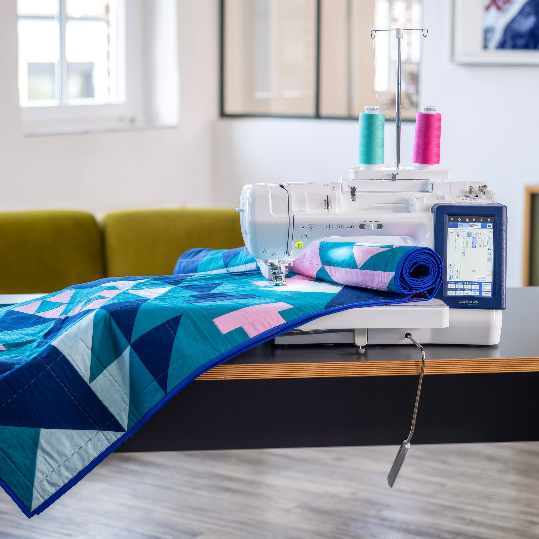 Brother BX1 machine with a digital screen and colorful thread spools sits on a wooden table, feeding a geometric quilt with blue, teal, and pink patterns.