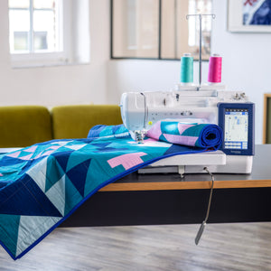 Brother BX1 machine with a digital screen and colorful thread spools sits on a wooden table, feeding a geometric quilt with blue, teal, and pink patterns.