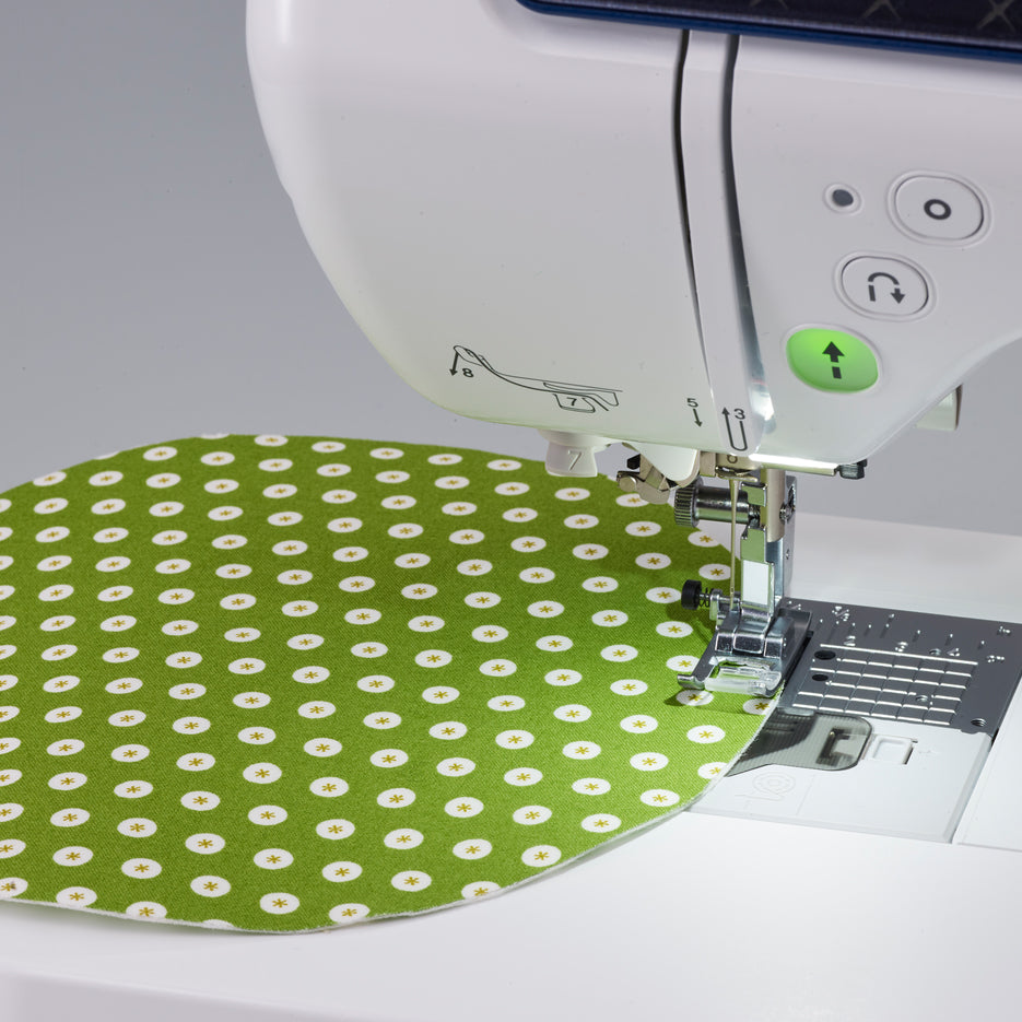 A close-up of a white sewing machine stitching a green fabric with a white daisy pattern under the needle and presser foot.