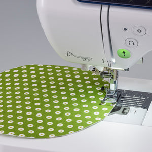 A close-up of a white sewing machine stitching a green fabric with a white daisy pattern under the needle and presser foot.