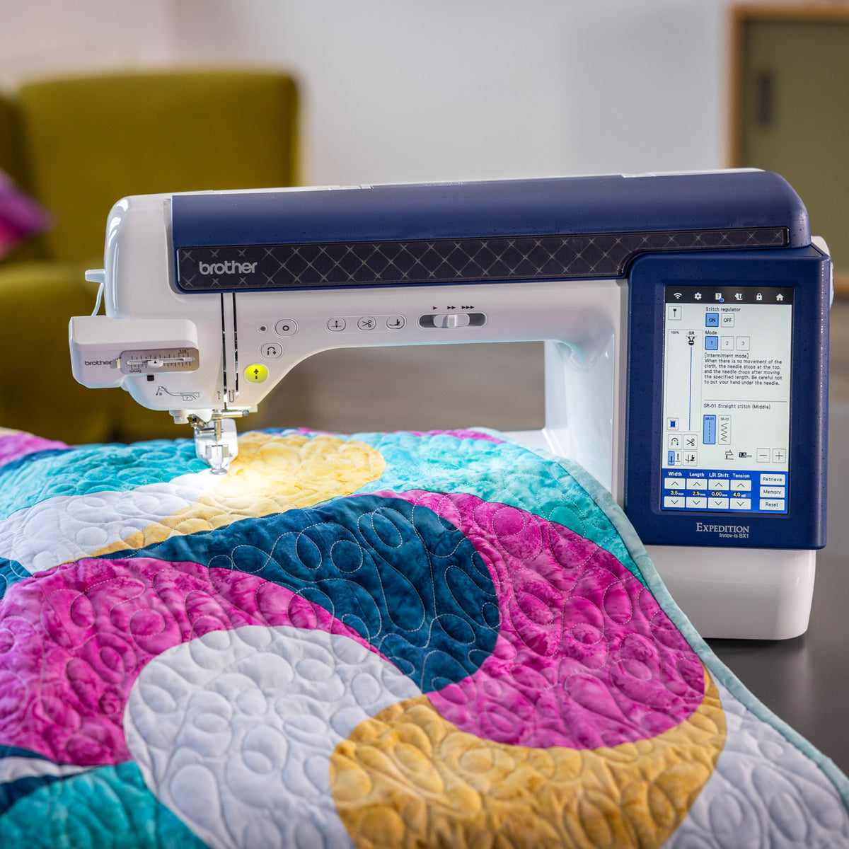 A Brother Quillcraft embroidery machine with a digital touchscreen display, shown actively stitching a colorful quilt with pink, blue, yellow, and white sections.