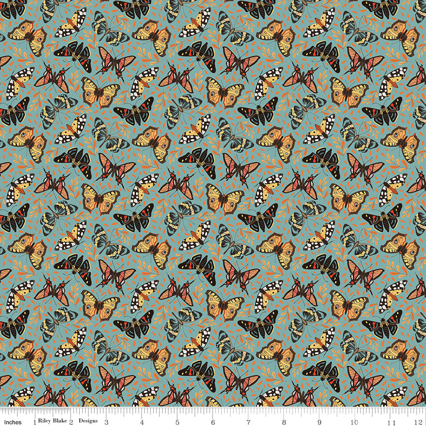 A repeating fabric pattern featuring orange, black, and white butterflies on a muted teal-blue background with small abstract orange scribbles. A ruler along the bottom edge shows scale.