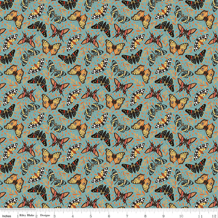 A repeating fabric pattern featuring orange, black, and white butterflies on a muted teal-blue background with small abstract orange scribbles. A ruler along the bottom edge shows scale.