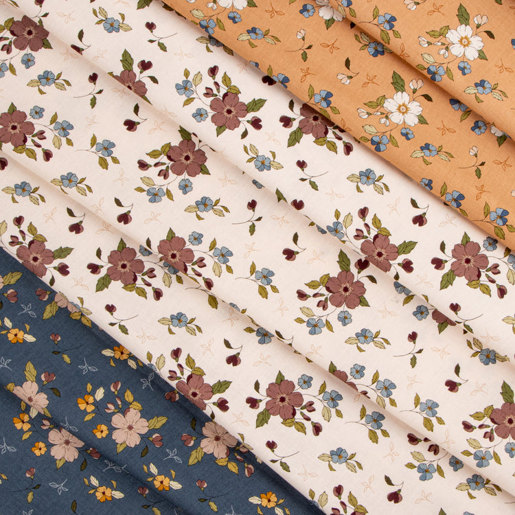 Folded fabrics with delicate floral prints arranged diagonally. The fabrics feature floral motifs in dusty pink, blue, yellow, and white on cream, peach, and navy backgrounds.