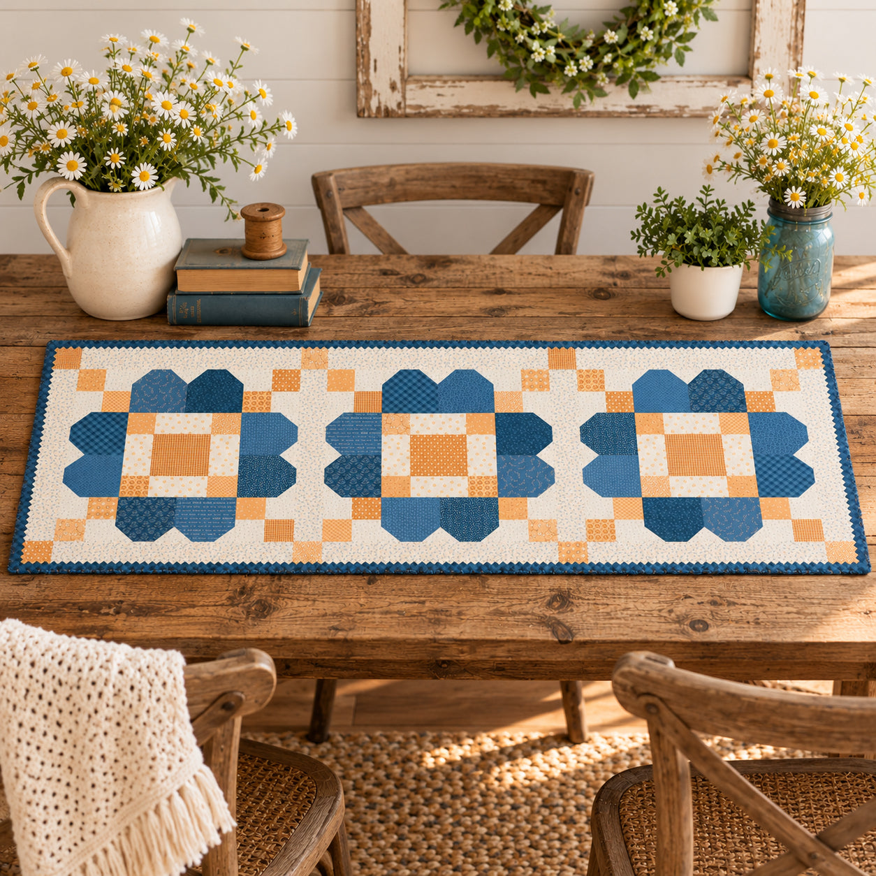A quilted table runner with a geometric block pattern in shades of blue, orange, and cream, styled on a wooden dining table with plates, glasses, and floral centerpieces.