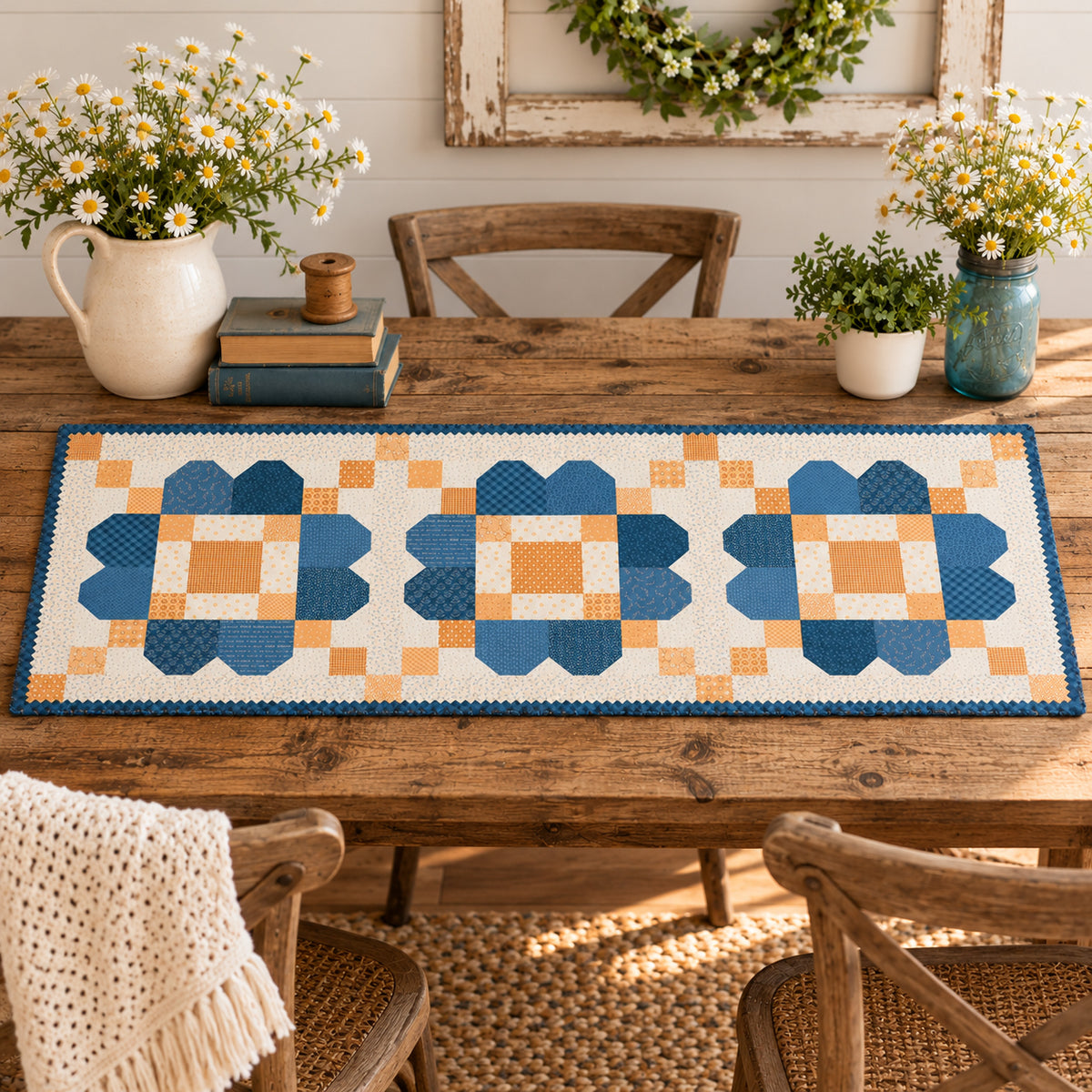A quilted table runner with a geometric block pattern in shades of blue, orange, and cream, styled on a wooden dining table with plates, glasses, and floral centerpieces.