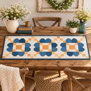 A quilted table runner with a geometric block pattern in shades of blue, orange, and cream, styled on a wooden dining table with plates, glasses, and floral centerpieces.