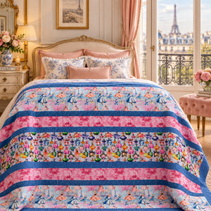 A colorful quilted comforter with pink and blue stripes and floral patterns is styled on a bed in a luxurious bedroom with a view of the Eiffel Tower.