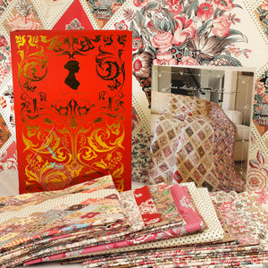 A styled product display of fabric and a quilt pattern. On the left is a red and gold decorative envelope or box, and in the center-right is a clear plastic-wrapped quilt pattern book showing a floral quilt design.
