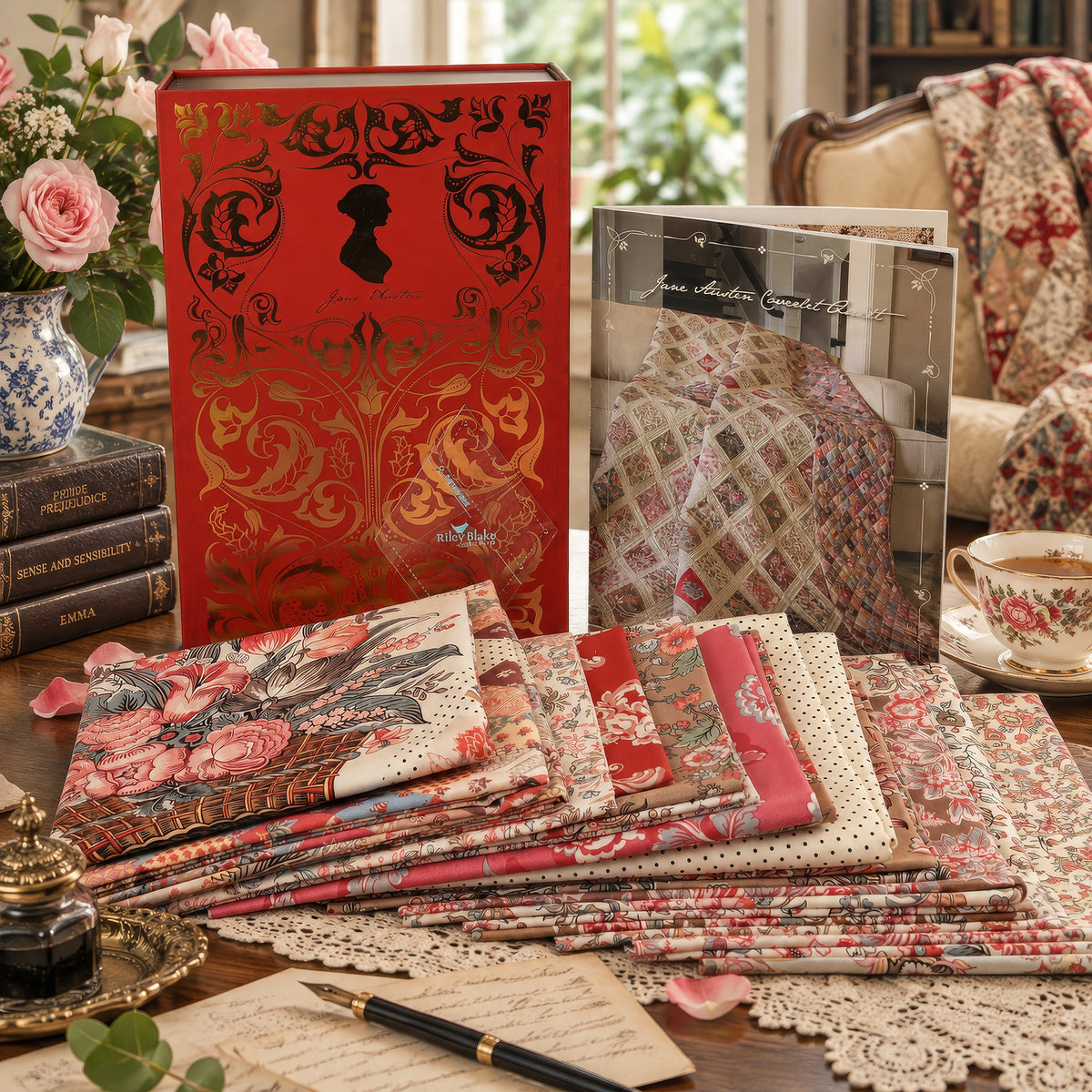 A styled tabletop display featuring a red decorative book, a fabric pattern booklet, and multiple folded quilting fabrics with floral and patchwork designs. The scene includes a teacup, lace doily, books, flowers, and a chair in a softly lit interior.