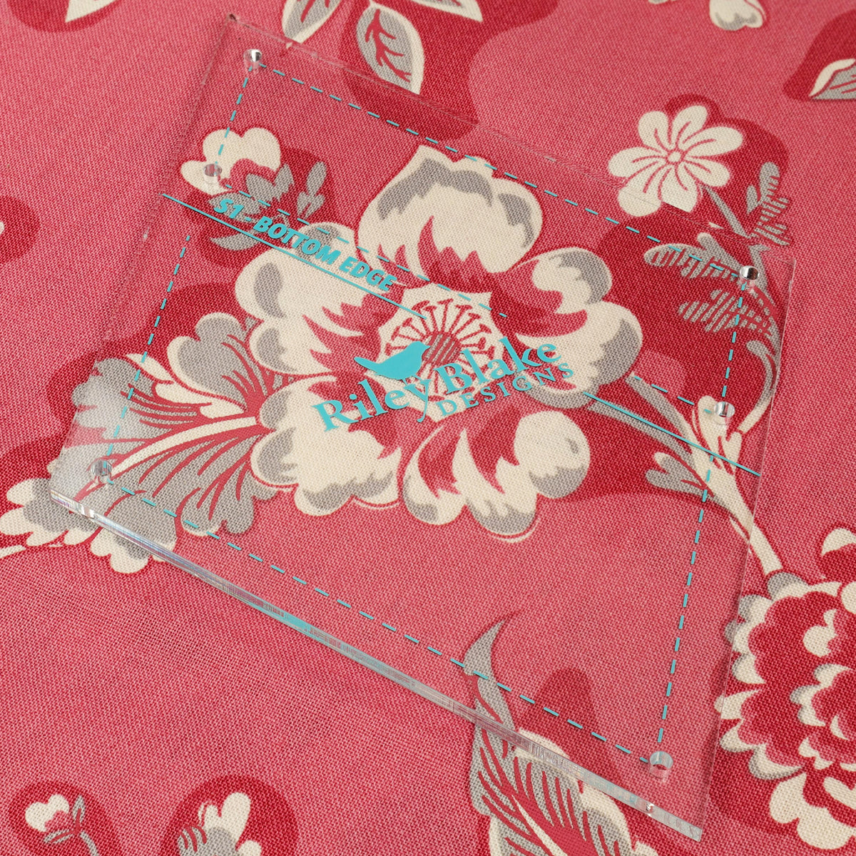A close-up of a pink floral fabric with a clear acrylic measuring square and blue dashed guide lines, showing a Balmain design logo.
