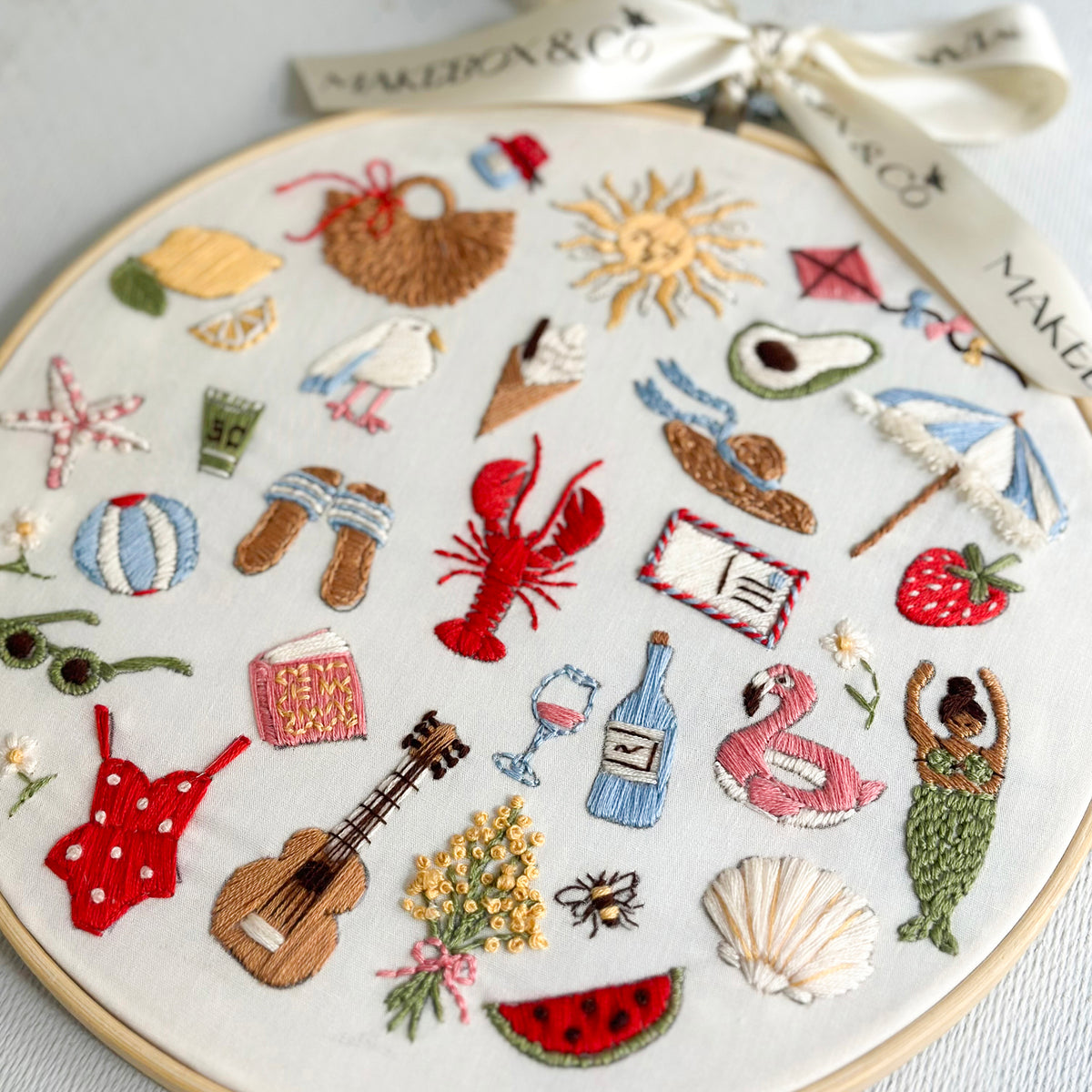 A circular embroidery hoop displays a colorful summer-themed embroidered patchwork design on a white fabric background, complete with a white satin ribbon.