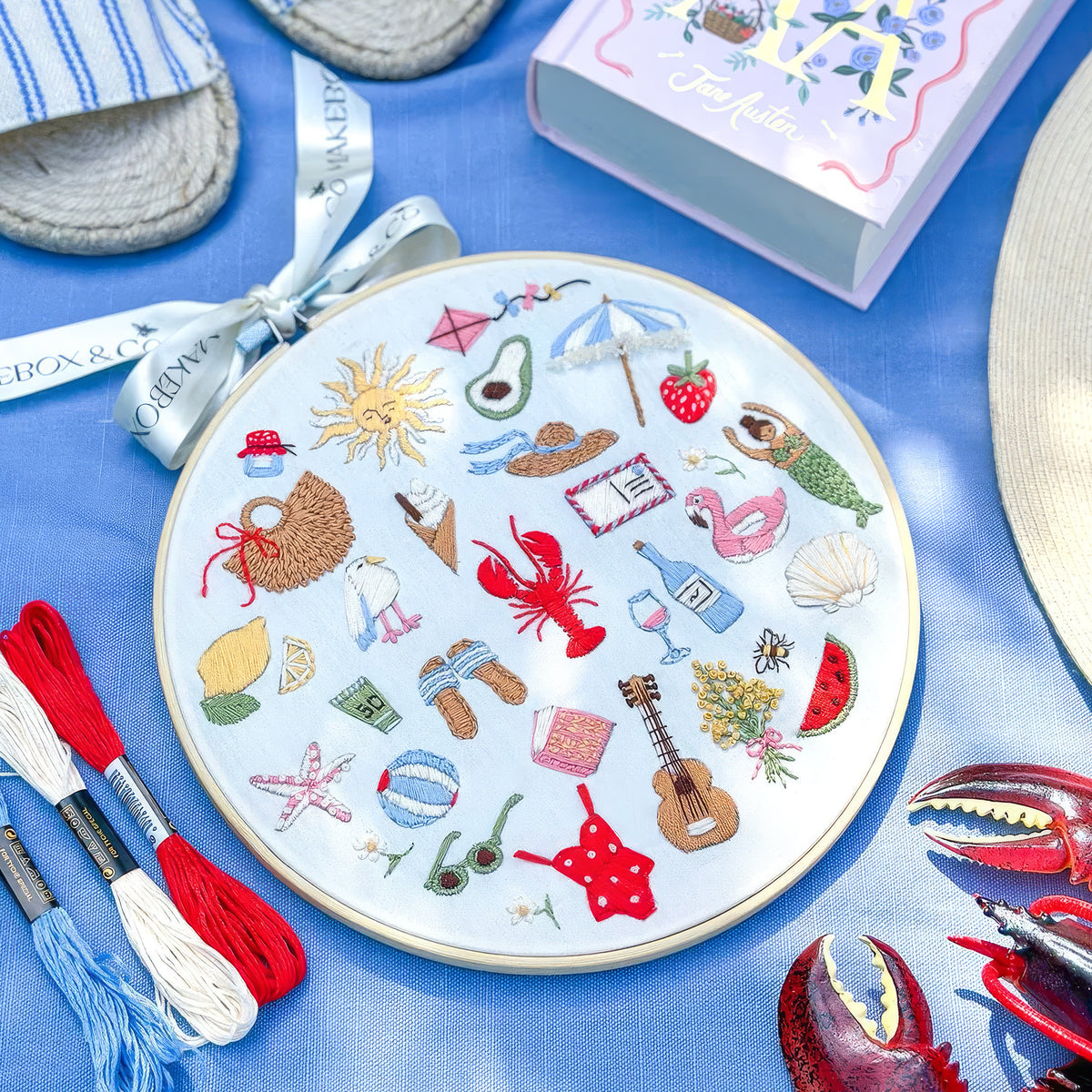 A circular embroidery hoop filled with colorful summer-themed embroidery on a light blue fabric background, surrounded by matching props including red and blue thread spools, a straw hat, a book, and a bracelet.