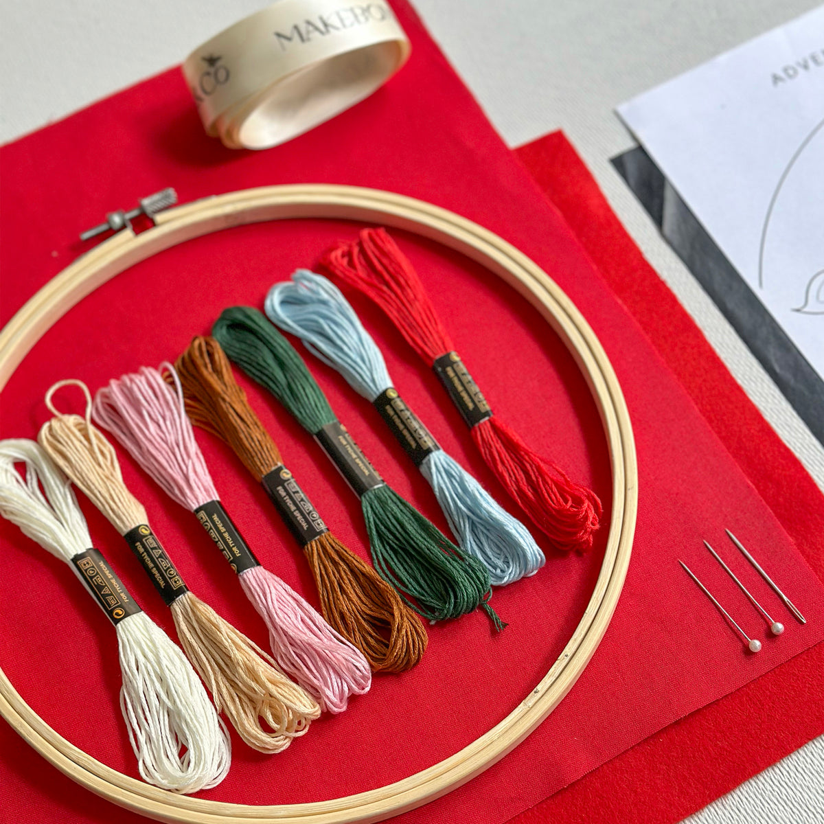 A circular embroidery hoop holding six skeins of embroidery floss in white, beige, pink, brown, light blue, and red, arranged on red fabric with pins and a small roll of tape nearby.