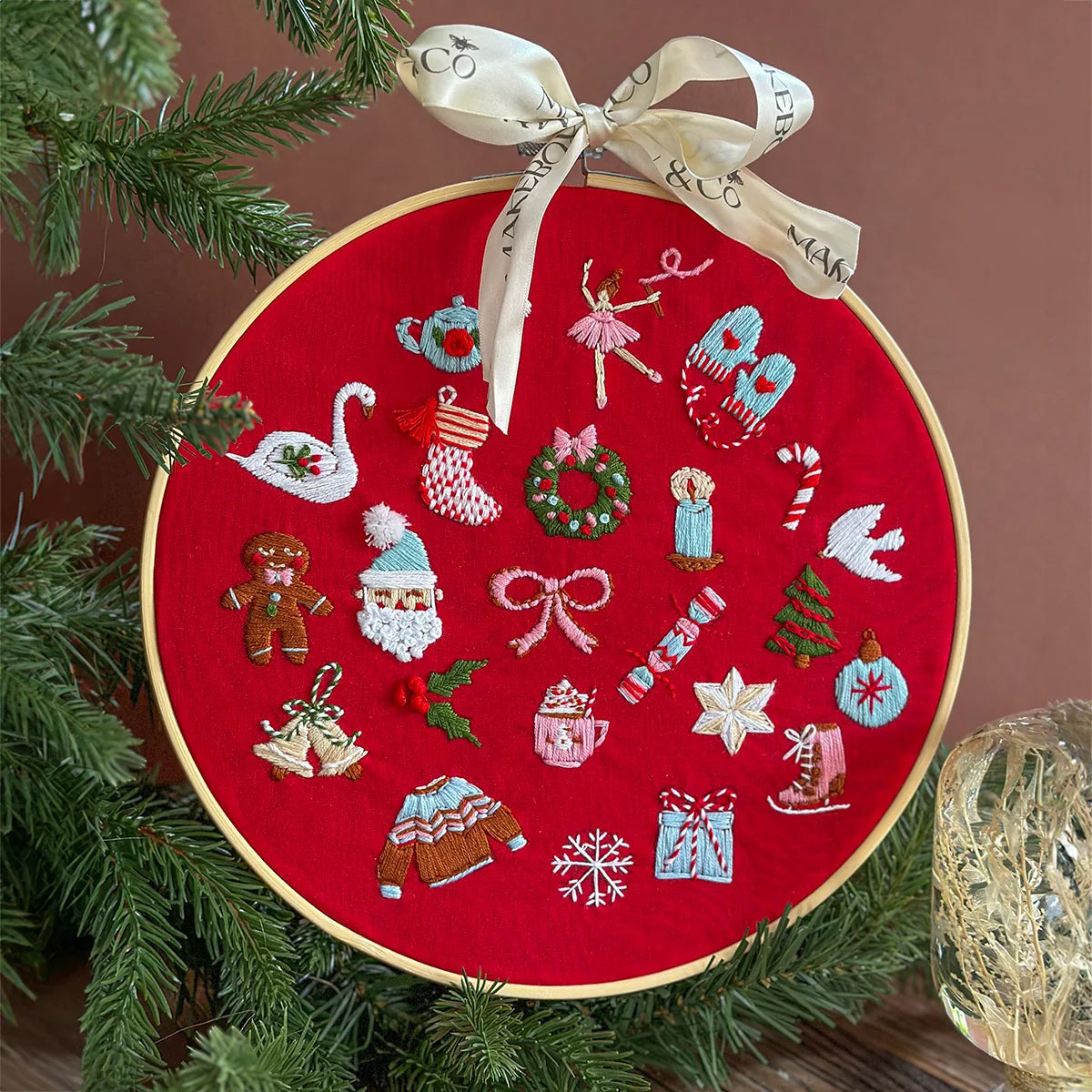 A round red fabric embroidery hoop ornament decorated with colorful Christmas-themed stitched motifs, hanging from a cream ribbon bow, styled with evergreen branches against a warm brown background.