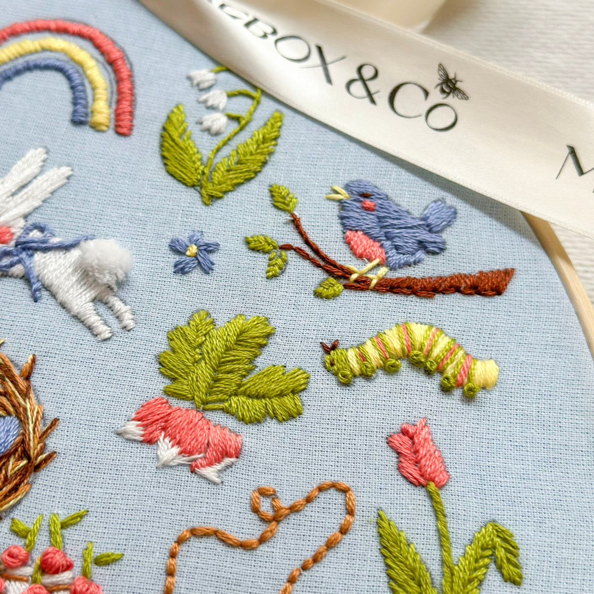 Close-up of a light blue embroidered fabric featuring colorful stitched animals and nature motifs, including a blue bird, green leaves, pink flowers, a rainbow, and a white ribbon with the text 'BOX & Co'.