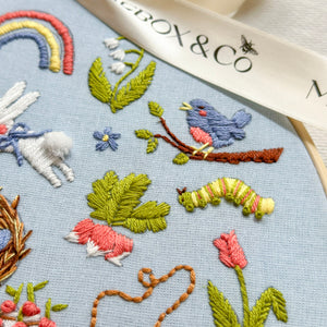 Close-up of a light blue embroidered fabric featuring colorful stitched animals and nature motifs, including a blue bird, green leaves, pink flowers, a rainbow, and a white ribbon with the text 'BOX & Co'.