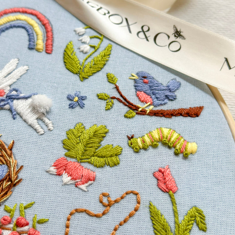 Close-up of a light blue embroidered fabric featuring colorful stitched animals and nature motifs, including a blue bird, green leaves, pink flowers, a rainbow, and a white ribbon with the text 'BOX & Co'.