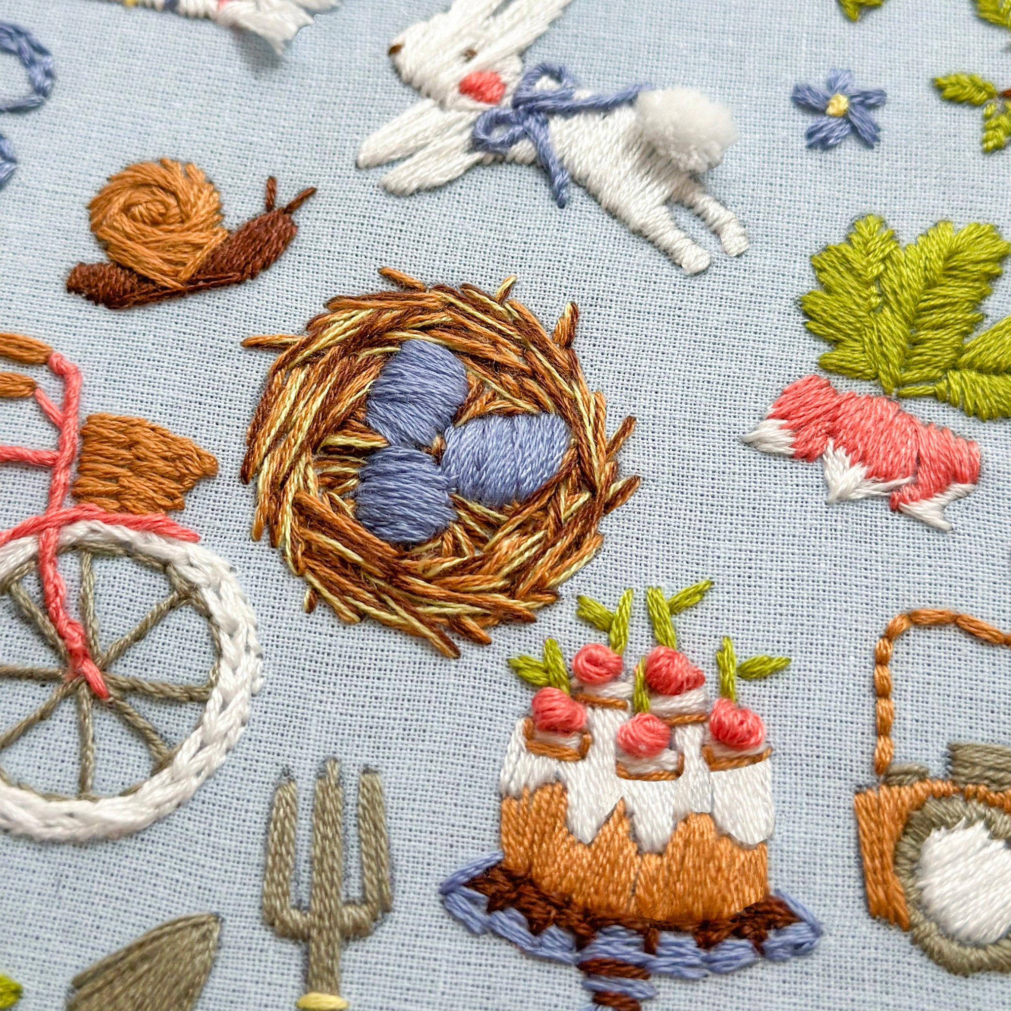 Close-up of a light blue embroidered fabric featuring whimsical folk-style motifs including a brown nest with blue eggs, a white rabbit with a blue bow, a snail, green leaves, pink flowers, and small embroidered cakes.