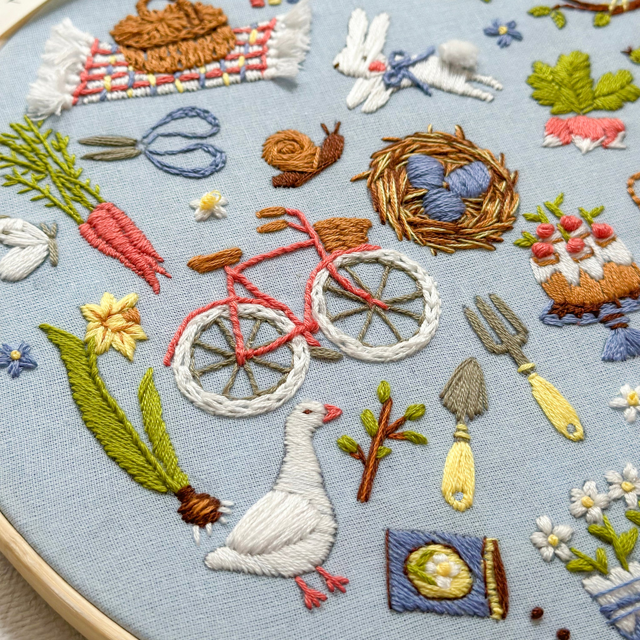 Close-up of a circular embroidery hoop showing a detailed multicolor embroidered scene on light blue fabric, featuring a red bicycle, white goose, carrots, flowers, birds, and gardening tools.
