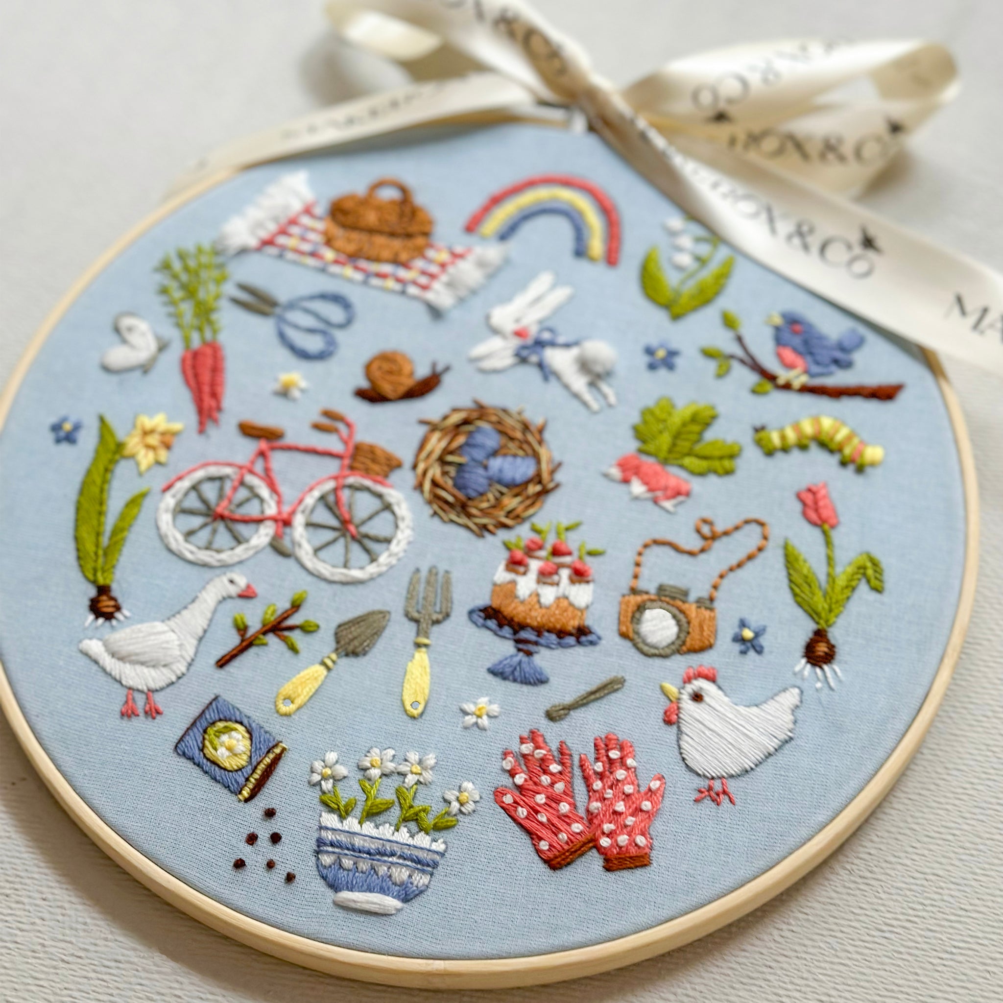 A circular embroidery hoop containing a detailed spring-themed embroidered design on light blue fabric, tied with a cream ribbon. The embroidery features various motifs including a rainbow, bicycle, flowers, birds, and a chicken.