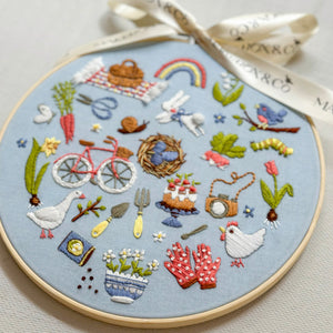 A circular embroidery hoop containing a detailed spring-themed embroidered design on light blue fabric, tied with a cream ribbon. The embroidery features various motifs including a rainbow, bicycle, flowers, birds, and a chicken.