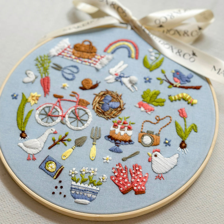 A circular embroidery hoop containing a detailed spring-themed embroidered design on light blue fabric, tied with a cream ribbon. The embroidery features various motifs including a rainbow, bicycle, flowers, birds, and a chicken.