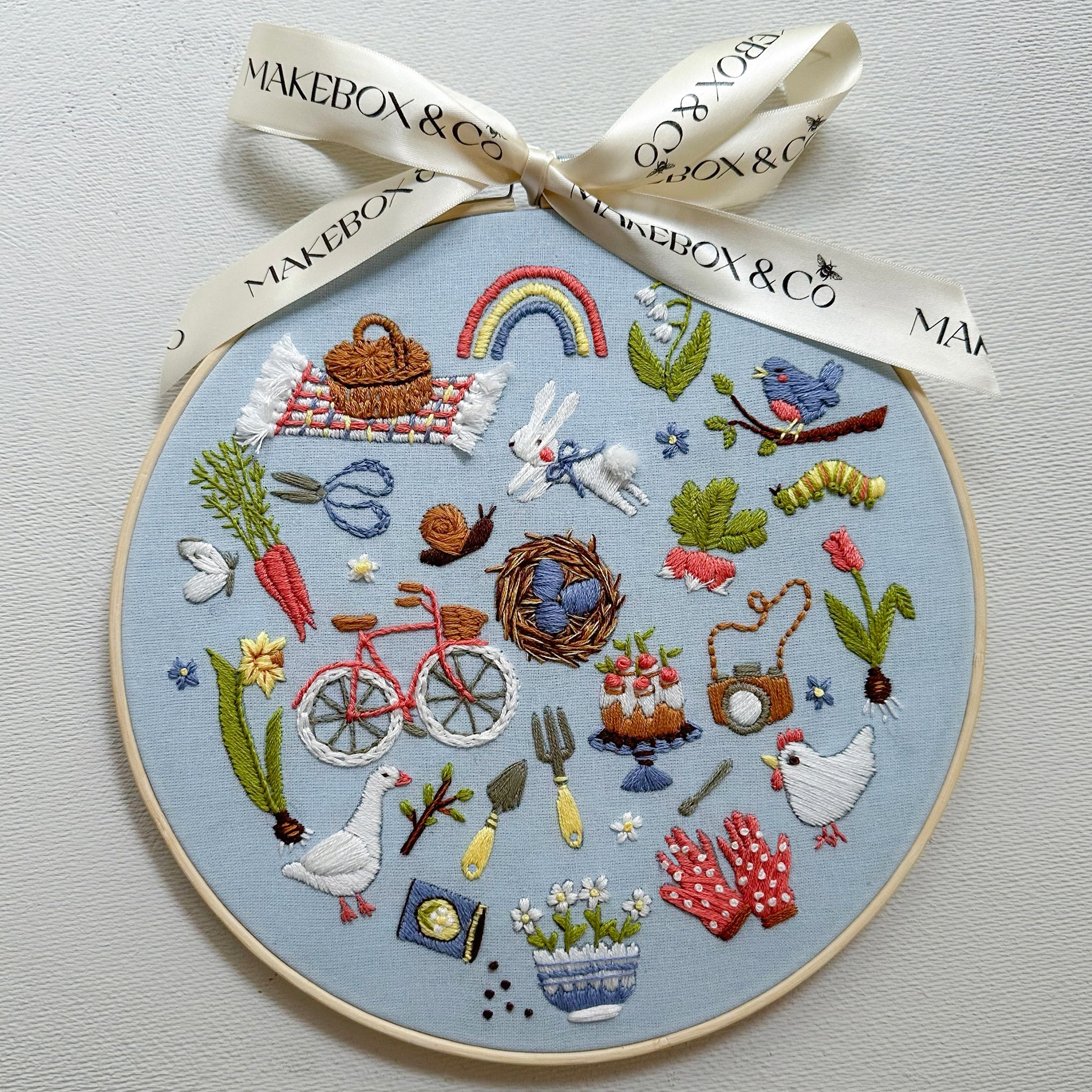 A circular embroidery hoop featuring a detailed Easter-themed embroidered design on a light blue fabric background, tied with a cream ribbon printed with the brand name 'MAKEBOX & CO'. The embroidery includes various spring motifs such as a rainbow, bunnies, carrots, a bicycle, a birdhouse, and a goose.