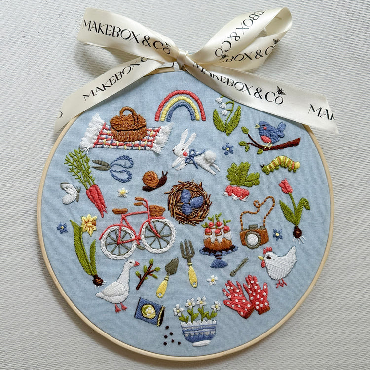 A circular embroidery hoop featuring a detailed Easter-themed embroidered design on a light blue fabric background, tied with a cream ribbon printed with the brand name 'MAKEBOX & CO'. The embroidery includes various spring motifs such as a rainbow, bunnies, carrots, a bicycle, a birdhouse, and a goose.