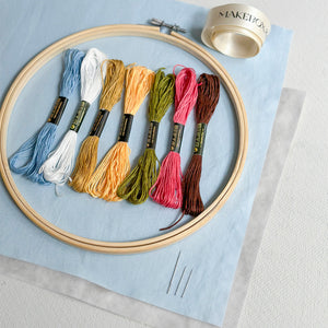 A flat-lay of embroidery supplies on a light blue fabric background, including a wooden hoop, a spool of thread, six skeins of embroidery floss in blue, white, yellow, olive green, pink, and brown, and two metal pins.