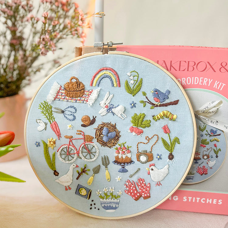 A round embroidery hoop displaying a colorful spring-themed stitched design on light blue fabric, with a pink 'Cakebox & Embroidery Kit' box behind it on a soft, blurred indoor background.