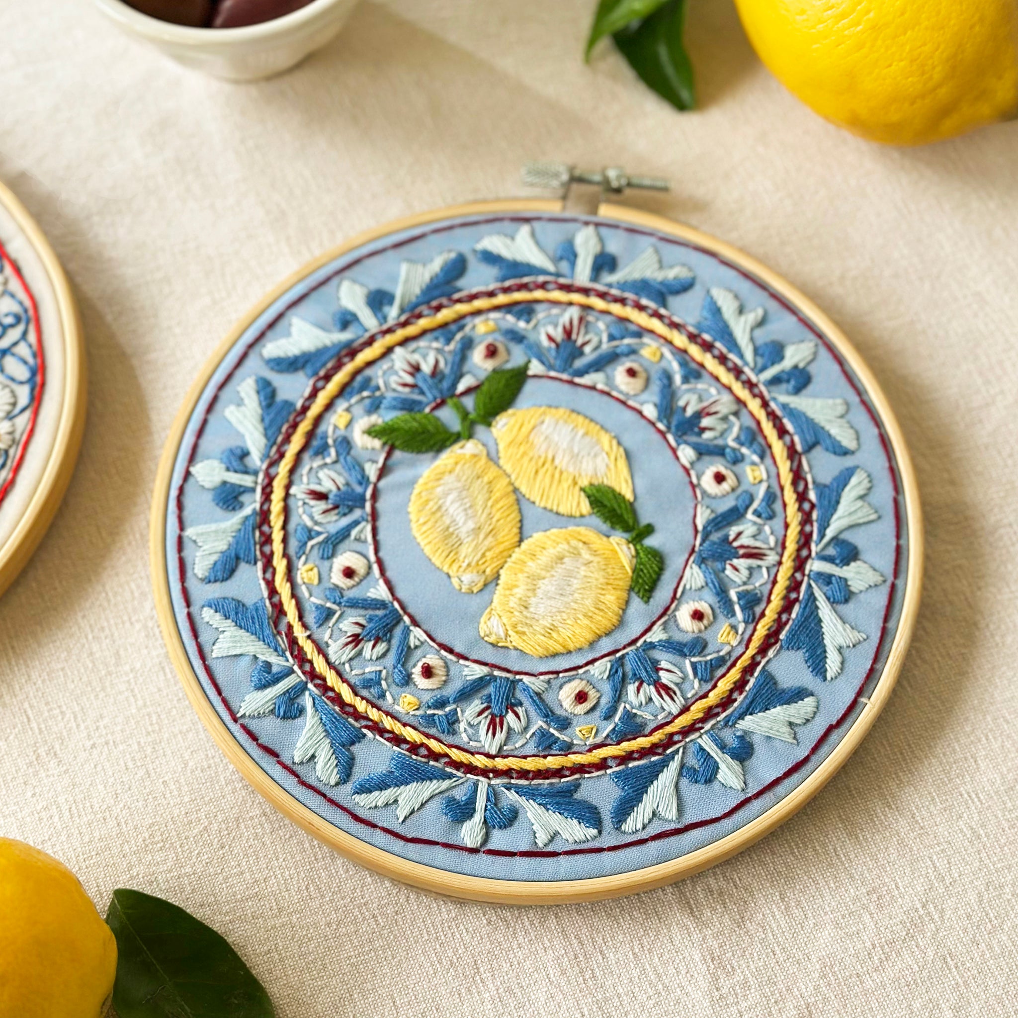 A circular embroidery hoop holding a detailed floral and lemon embroidery design on light blue fabric, styled on a beige textured surface with lemons nearby.