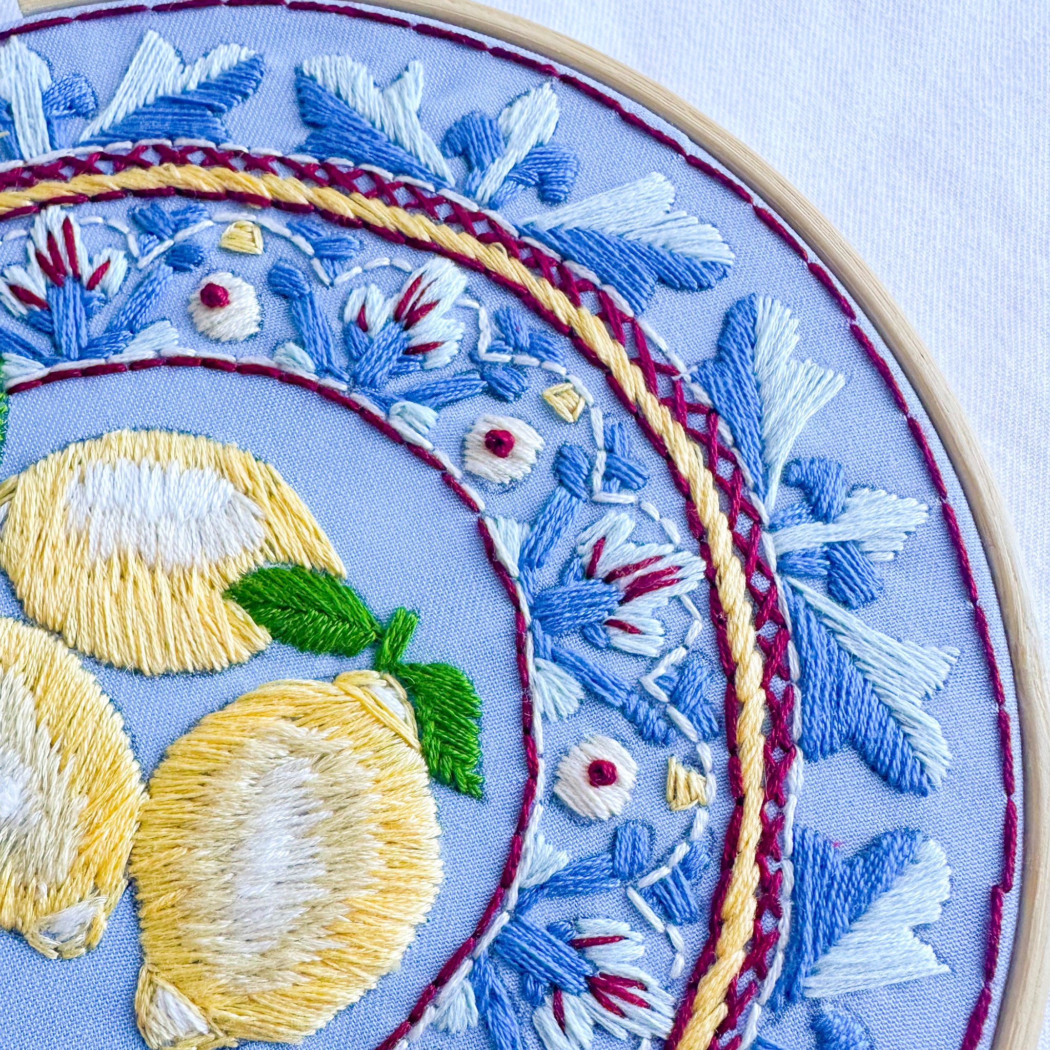Close-up of a circular embroidery hoop showing detailed floral and lemon embroidery on a light blue fabric background.