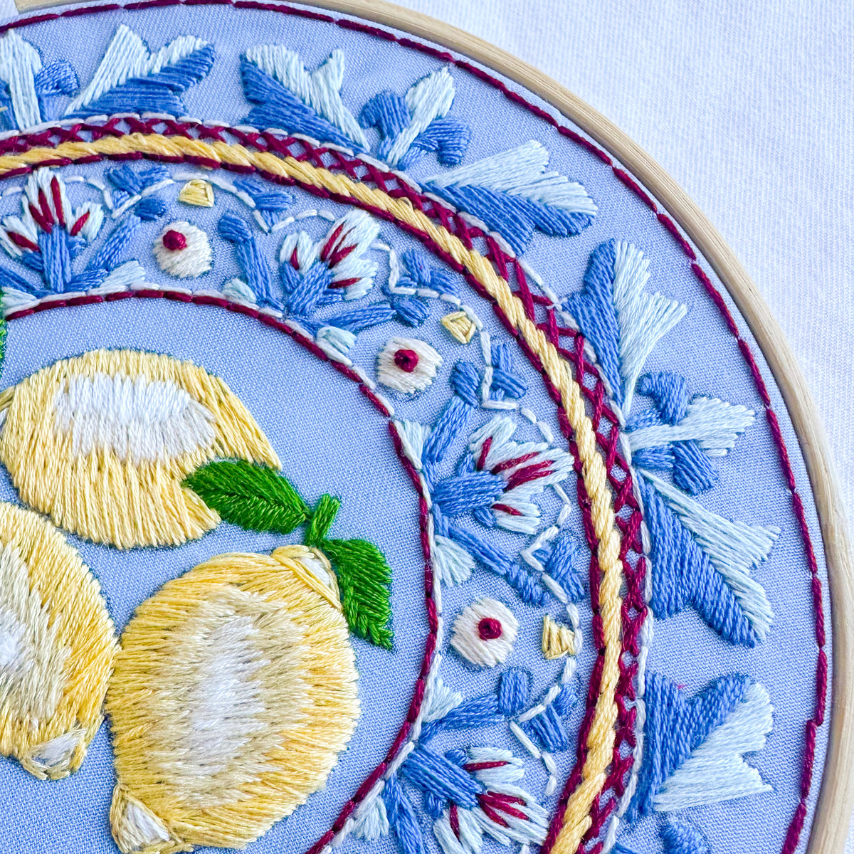Close-up of a circular embroidery hoop showing detailed floral and lemon embroidery on a light blue fabric background.
