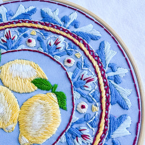 Close-up of a circular embroidery hoop showing detailed floral and lemon embroidery on a light blue fabric background.
