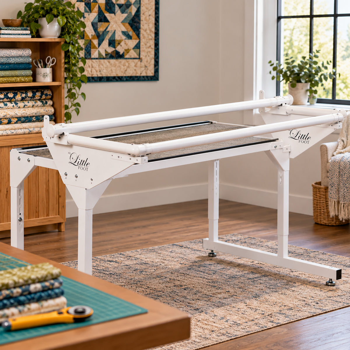 Little Foot quilting frame set up in a bright sewing room with wood floors, a quilted wall hanging, plants, and fabric stacks nearby.