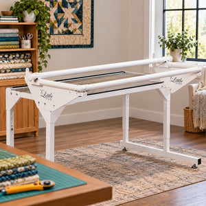 Little Foot quilting frame set up in a bright sewing room with wood floors, a quilted wall hanging, plants, and fabric stacks nearby.