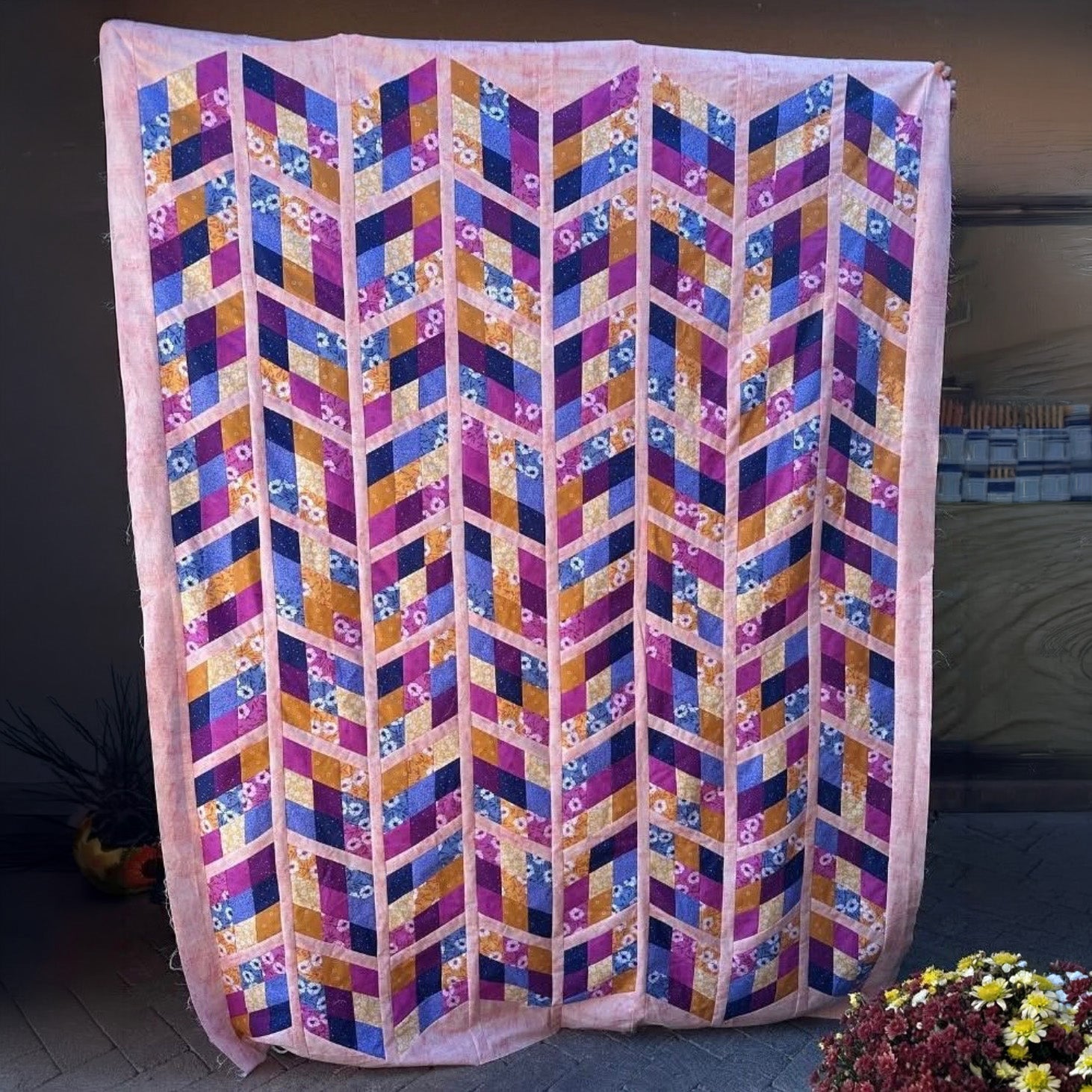 A large quilted textile panel or wall hanging with a geometric chevron pattern in pink, purple, blue, orange, and gold, displayed outdoors against a dark wall with flowers at the bottom.
