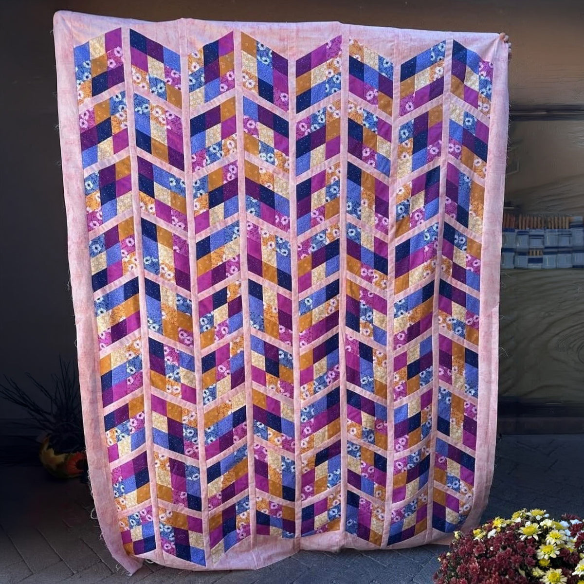 A large quilted textile panel or wall hanging with a geometric chevron pattern in pink, purple, blue, orange, and gold, displayed outdoors against a dark wall with flowers at the bottom.