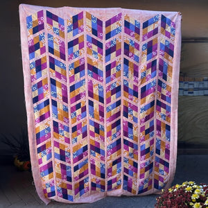 A large quilted textile panel or wall hanging with a geometric chevron pattern in pink, purple, blue, orange, and gold, displayed outdoors against a dark wall with flowers at the bottom.