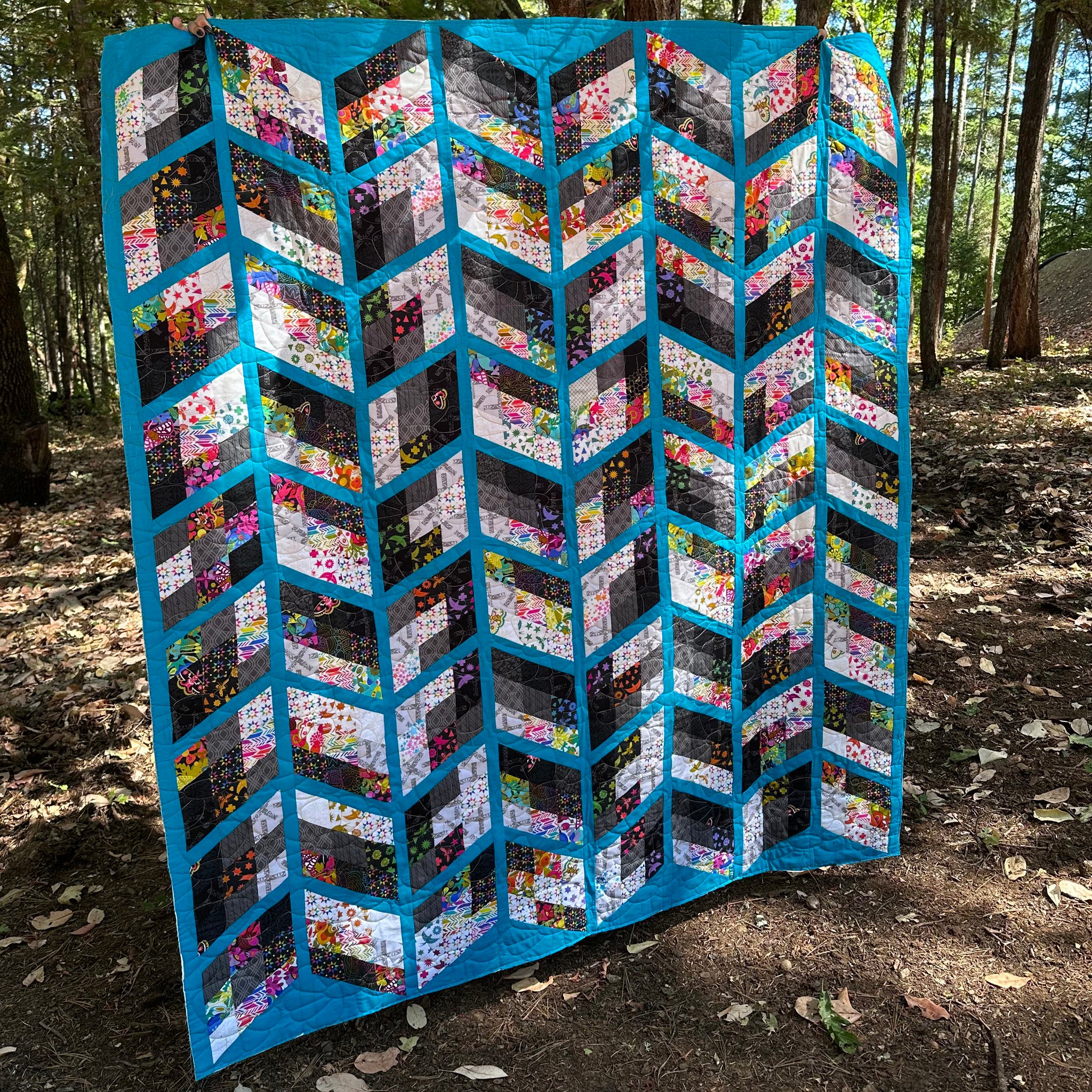 A large handmade quilt with a geometric chevron pattern, displayed outdoors in a wooded area. The quilt features bright turquoise blue borders and alternating rows of floral and abstract patterned fabric panels in black, white, pink, yellow, and green.