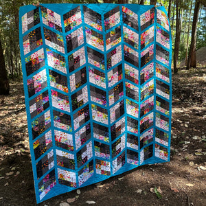 A large handmade quilt with a geometric chevron pattern, displayed outdoors in a wooded area. The quilt features bright turquoise blue borders and alternating rows of floral and abstract patterned fabric panels in black, white, pink, yellow, and green.
