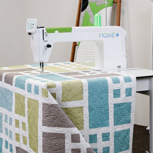 Moxie ST sewing machine is shown quilting a large patchwork quilt with geometric blocks in blue, sage green, taupe, and white. The sewing machine features a digital controller on the arm and the Moxie brand logo.